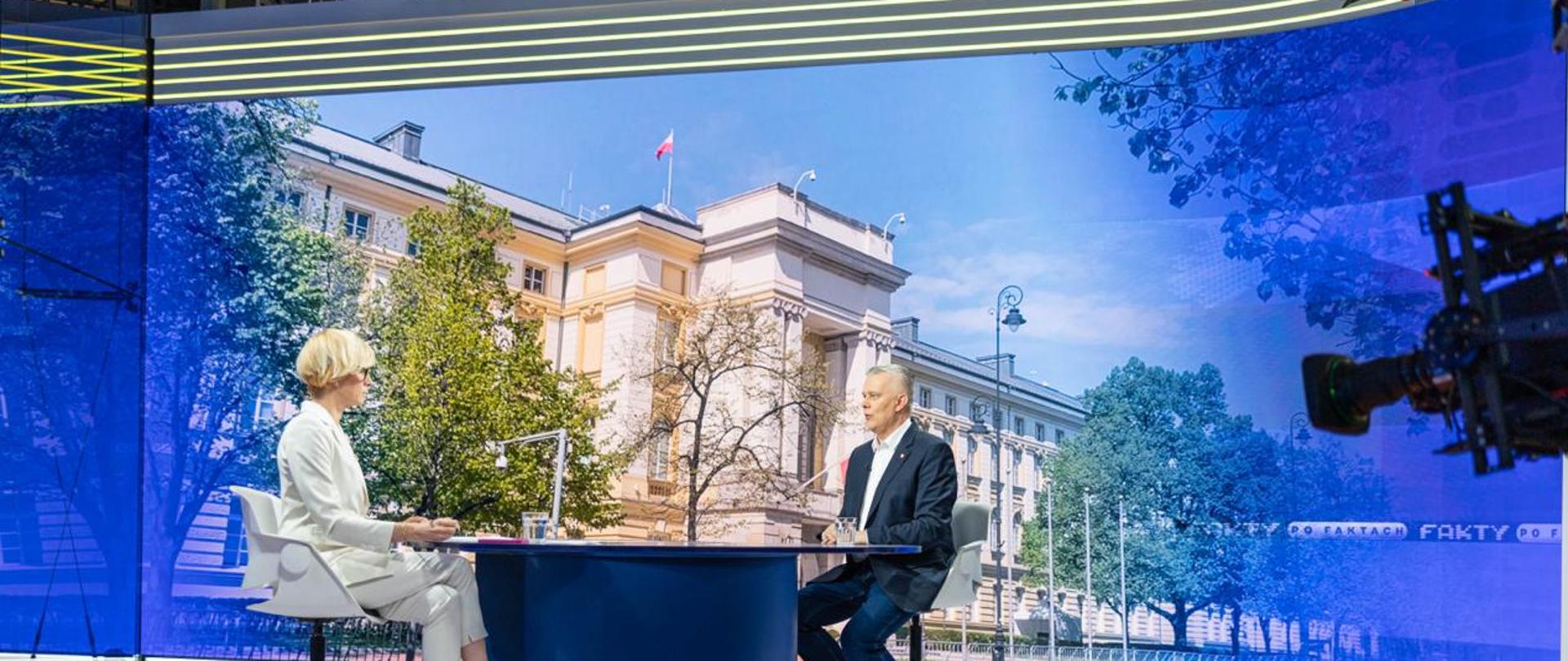 Na zdjęciu studio telewizyjne. Przy niebieskim stole, na białych krzesłach siedzą dwie osoby - po prawej minister Tomasz Siemoniak, po lewej - redaktor Anita Werner. Na stole dwie szklanki z wodą oraz materiały. W tle zdjęcie frontu budynku Kancelarii Prezesa Rady Ministrów. Z prawej strony zdjęcia widać fragment kamery.