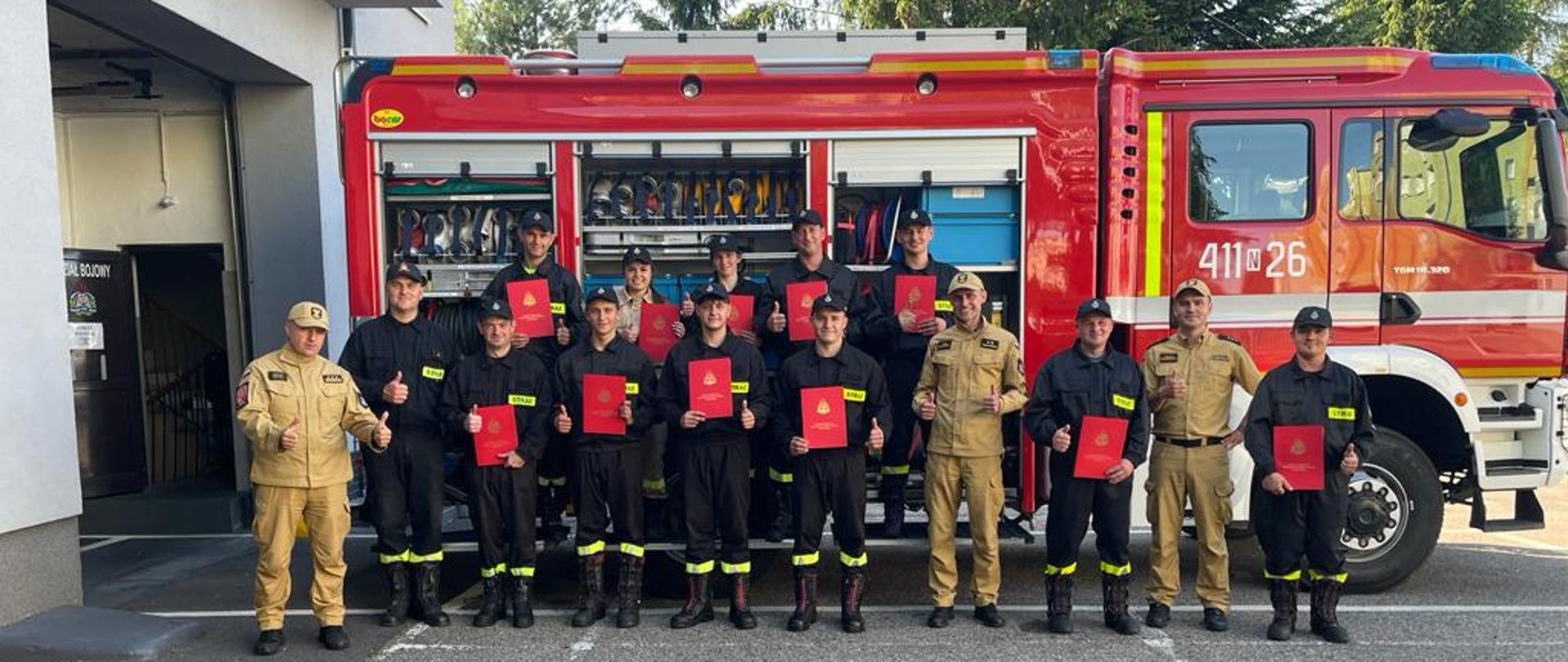 Na zdjęciu pozują na tle samochodu strażackiego, uczestnicy kursu, w rekach trzymają czerwone dyplomy oraz pracownicy komendy.