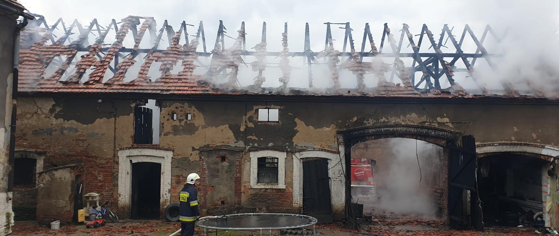 Obraz przedstawia budynek stodoły po pożarze. Widoczna spalona więźba dachowa. Z wnętrza wydobywa się dym i para wodna.