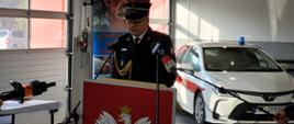 Strażak w mundurze galowym podczas przemówienia w pomieszczeniu garażowym. W tle biały samochód.