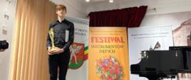 na zdjęciu na tle plaktów z festiwalu Instrumentów dętych uczeń w czarnym ubraniu ze złotą statuetką w rękach