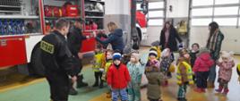 Pomieszczenie garaży jednostki ratowniczo gaśniczej w Sępólnie Krajeńskim, przy samochodzie ratowniczo gaśniczym z podniesionymi roletami stoi dwóch strażaków w ubraniach koszarowych oraz kilkanaścioro przedszkolaków wraz z opiekunkami