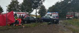 Wypadek na trasie Kościan - Nowy Lubosz. Dwa rozbite samochody osobowe. Strażacy i ratownicy medyczni udzielają pomocy osobom poszkodowanym w zderzeniu pojazdów. w tle karetki i samochody strażackie.