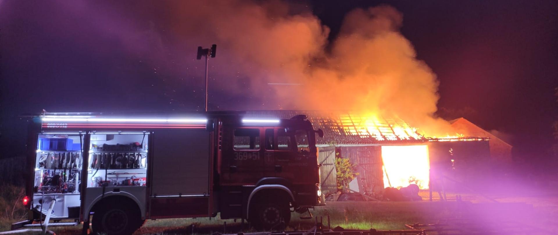 Zdjęcie przedstawia pożar budynku gospodarczego, płomienie wychodzą ponad dach budynku, w tle samochód strażacki