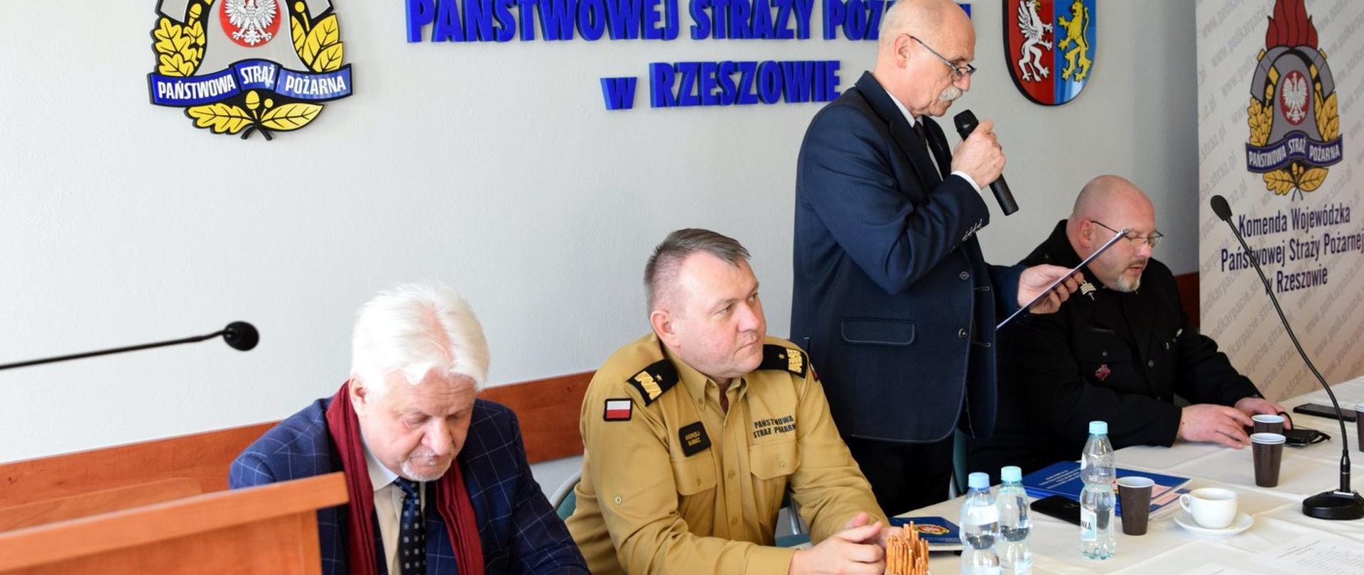 djęcie zrobione w sali konferencyjnej. Przy stole jest czterech uczestników spotkania. Jeden z nich stoi i przemawia do mikrofonu. Wśród osób siedzących jest oficer państwowej straży pożarnej w stopniu generała. 
