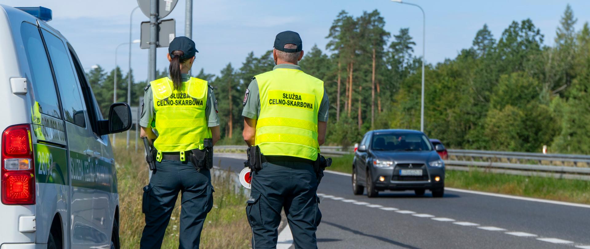 Funkcjonariusze stoją przy radiowozie i patrzą na drogę, którą jedzie samochód