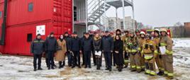 Na tle trenażera pozuje do zdjęcia grupa strażaków oraz cywilów, w tym trzy kobiety