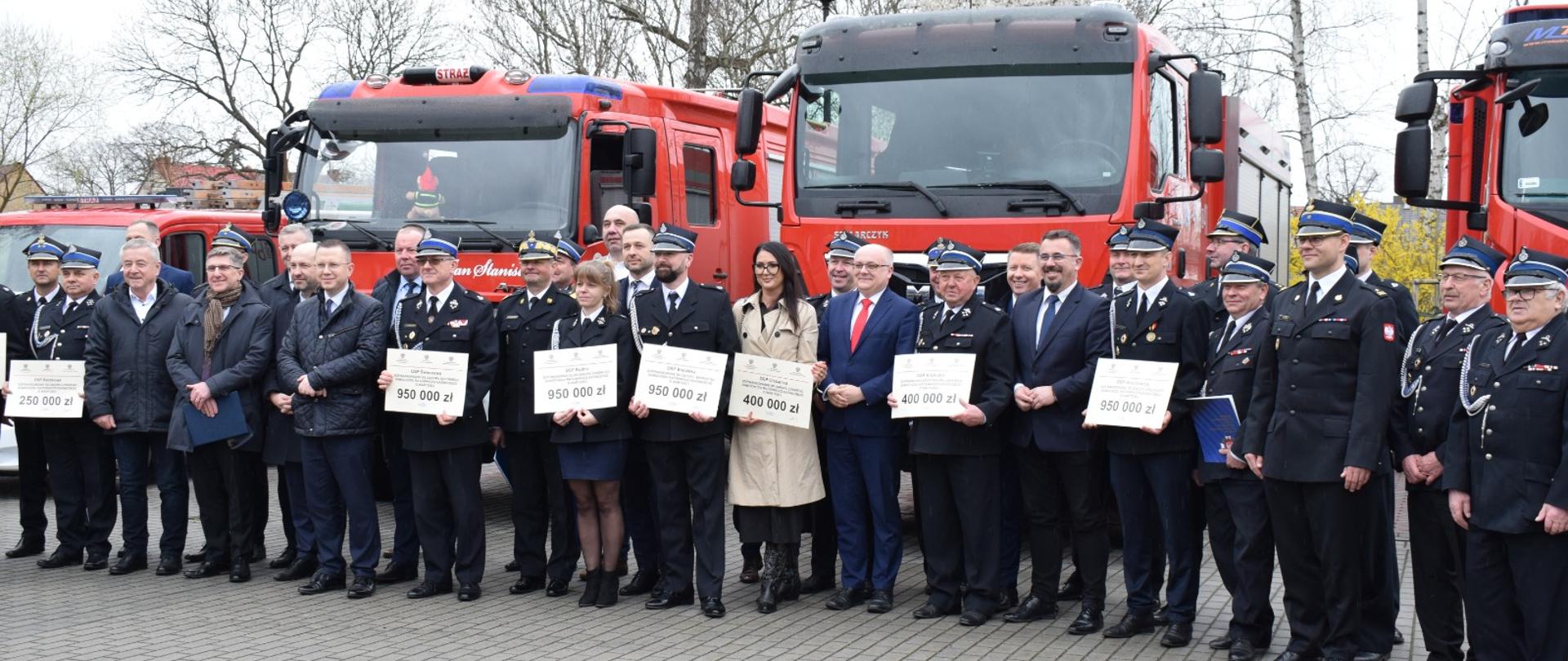 Zdjęcie grupowe osób stojących na tle czerwonych samochodów pożarniczych. Część osób trzyma otrzymane promesy