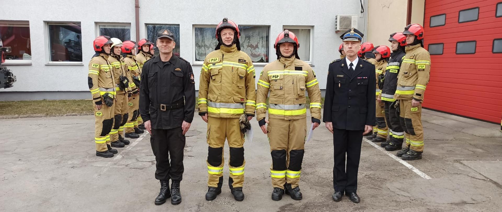 Zdjęcie przedstawia plac przed czerwonymi bramami garażowymi Jednostki Ratowniczo-Gaśniczej w Świdwinie. Przed bramą stoi zmiana służbowa w piaskowych ubraniach specjalnych z czerwonymi hełmami - stoją wzdłuż namalowanej białej linii. Na pierwszym planie znajduje się Komendant Powiatowy PSP w Świdwinie, dowódca JRG ubrani w czarne mundury. Obok nich stoją dwóch wyróżnionych strażaków ubrani w piaskowy mundur i czerwony hełm. W ręku trzymają dokumenty. Panowie się uśmiechają.