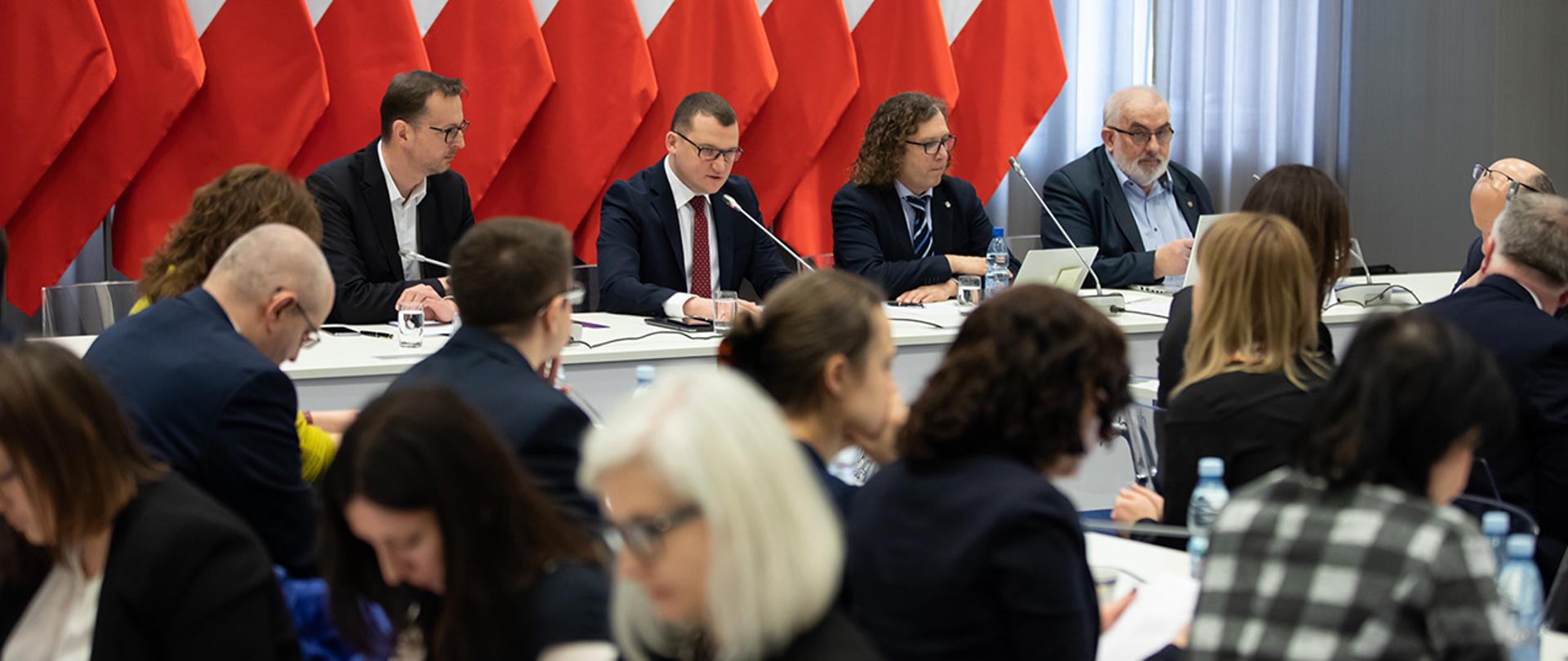 Na zdjęciu widać siedzących za stołem wiceministra Pawła Szefernakera współprzewodniczącego KWRiST, Jacka Karnowskiego współprzewodniczącego, Andrzeja Porawskiego Sekretarz strony samorządowej KWRiST i Szymona Wróbla zastępcę dyrektora Departamentu Administracji Publicznej MSWiA Sekretarza strony rządowej KWRiST. Widać również uczestników spotkania siedzących na sali zza których zostało wykonane zdjęcie. W tle widać rząd flag Polski.