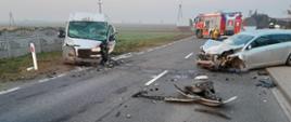 Na zdjęciu widać dwa rozbite samochody, srebrny osobowy i biały dostawczy. Oba pojazdy stoją po dwóch stronach drogi i znajdują się częściowo na jezdni a częściowo na poboczu. Na jezdni leżą porozrzucane elementy pojazdów. W tle widać stojący na drodze czerwony samochód straży pożarnej oraz strażaków. W oddali widać zabudowania, krzewy, słupy energetyczne oraz pola. Jest widno.