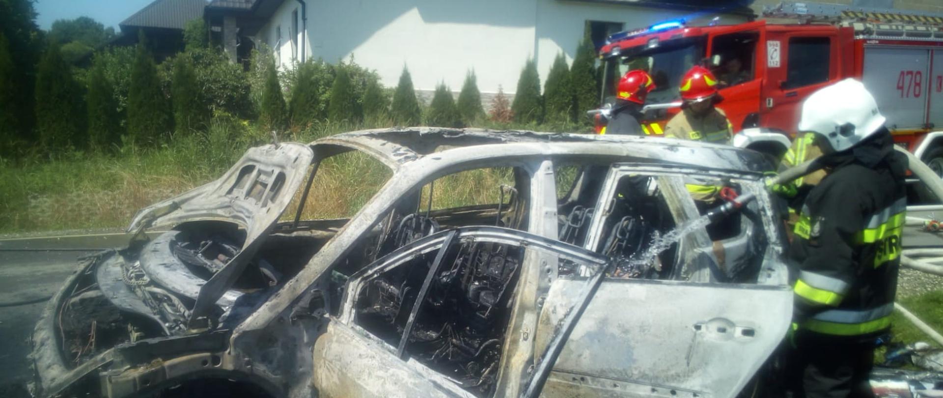 Na zdjęciu widoczny jest spalony samochód osobowy. Strażacy sprawdzają i dogaszają zarzewia ognia w pojeździe. W tle znajdują się zabudowania miejscowości. Z prawej strony na jezdni stoi samochód strażacki z jednostki OSP.