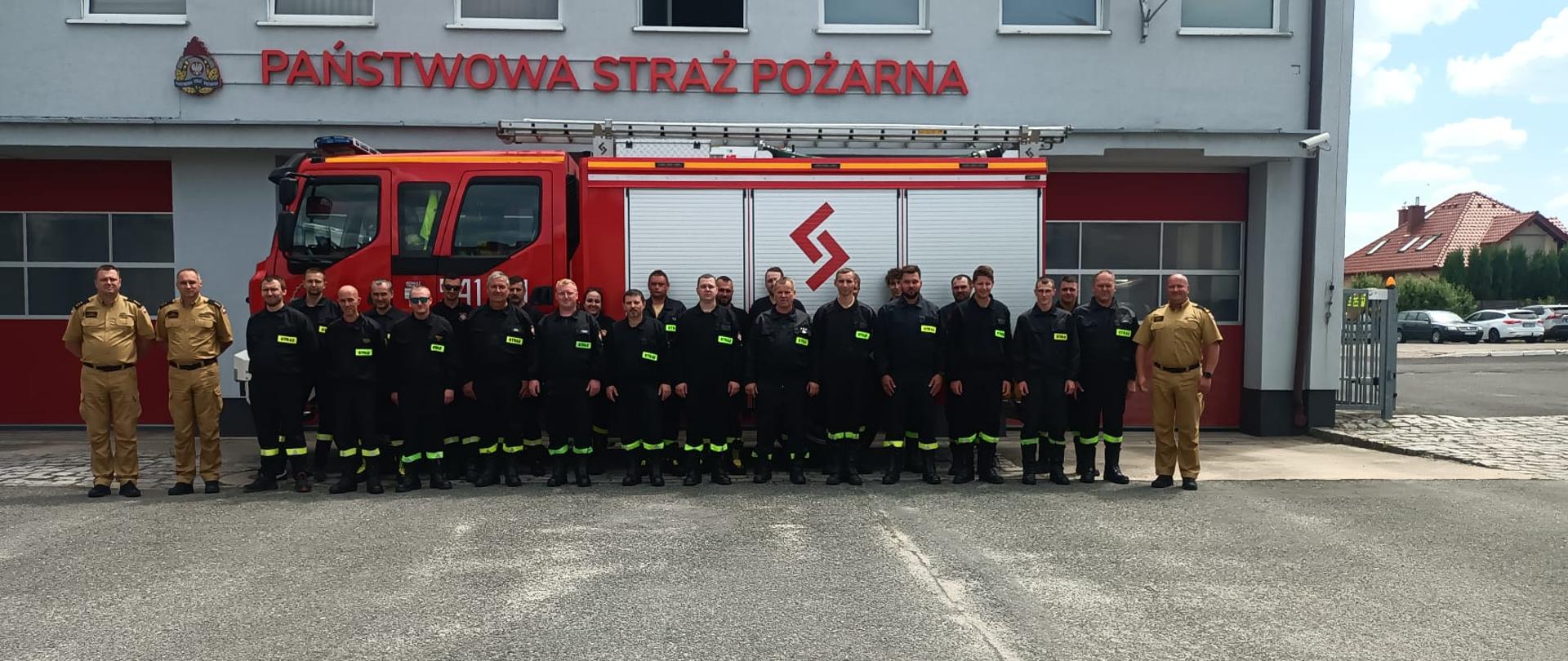Strażacy stojący na tle samochodu pożarniczego i komendy