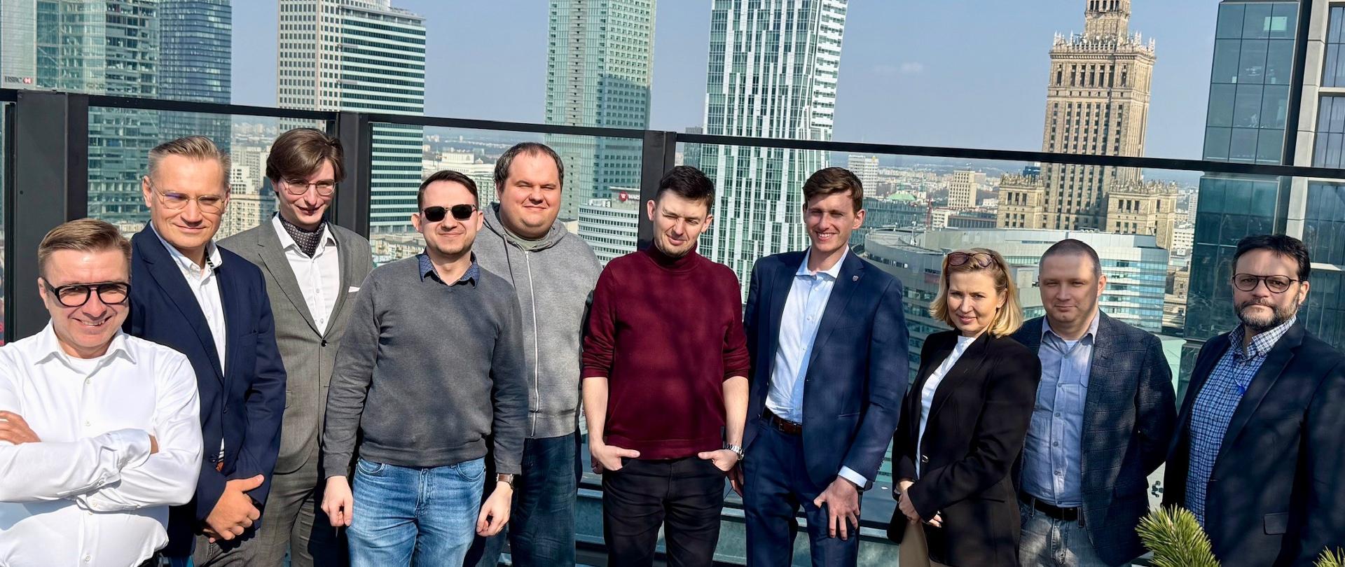 Zdjęcie grupowe dziesięciu osób pozujących na nowoczesnym, nasłonecznionym tarasie widokowym w Warszawie. W tle rozciąga się panorama miasta pod błękitnym niebem, z wyraźnie widocznym po prawej stronie Pałacem Kultury i Nauki oraz szklanymi wieżowcami w centrum. Uczestnicy to ośmiu mężczyzn i dwie kobiety, ubrani w stroje biznesowe oraz casualowe (marynarki, koszule, swetry, dżinsy). Większość z nich uśmiecha się patrząc w obiektyw.