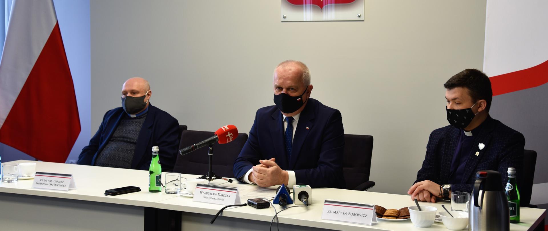 Wojewoda Lubuski Władysław Dajczak, ks. dr hab. Dariusz Śmierzchalski-Wachocz, ks. Marcin Bobowicz w czasie konferencji prasowej. Siedzą przy stole prezydialnym, w tle flagi, godło i baner Lubuskiego Urzędu Wojewódzkiego. Wojewoda Lubuski wypowiada się do mediów.