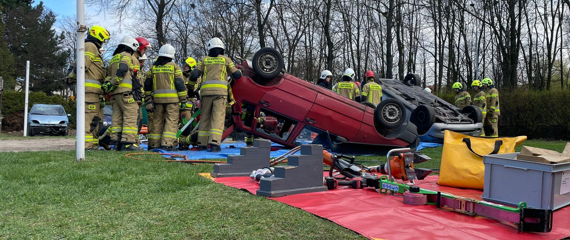 Strażacy ubrani w ubrania specjalne stoją wokół dwóch aut przewróconych na dach, obok widoczny rozłożony sprzęt hydrauliczny