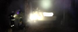 Pora nocna, dym przesłania budynek w którym znajduje się samochód osobowy w białym kolorze. W budynku w dymie mało widoczni strażacy. Po lewej stronie budynku dwóch strażaków z aparatami ochrony dróg oddechowych.
