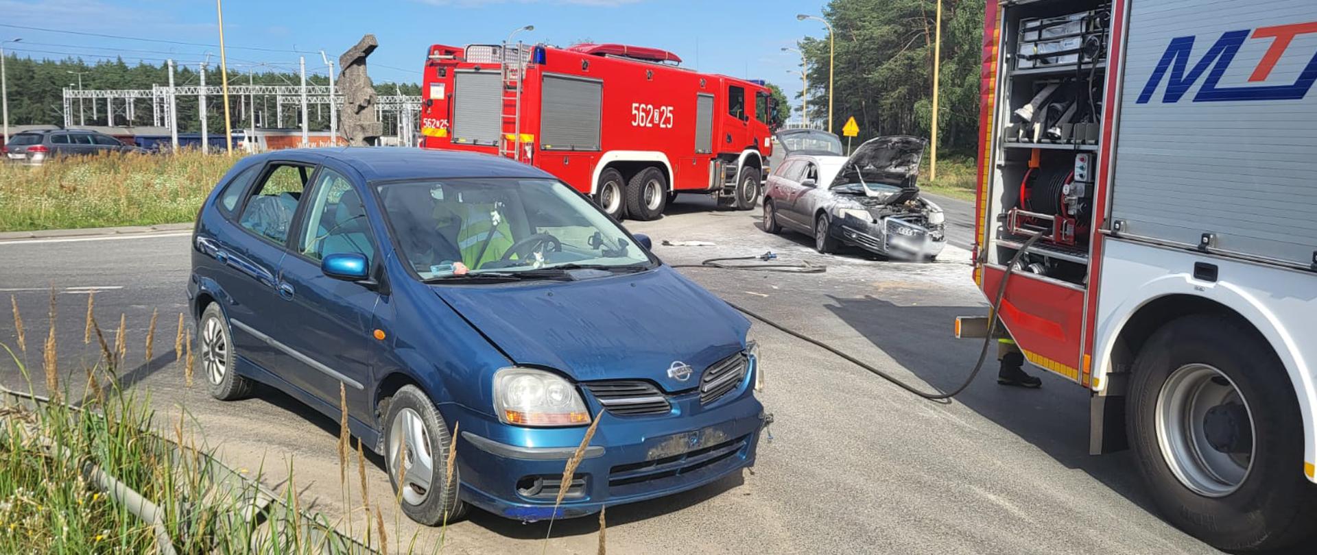 Zdjęcie przedstawia samochód uszkodzony w wyniku zderzenia samochód osobowy marki nissan koloru niebieskiego oraz samochód osobowy marki Ausi z otwartą maską oraz widocznym osadem z proszku gaśniczego. W tle samochody pożarnicze koloru czerwonego, rozwinięta linia gaśnicza od szybkiego natarcia. Samochody stoją na skrzyżowaniu na drodze. 