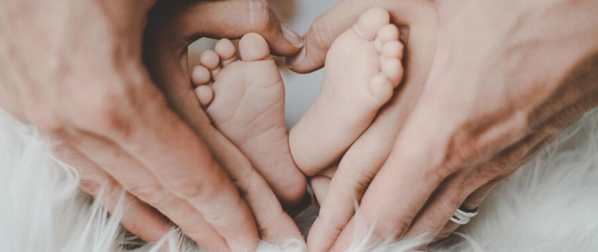 Na zdjęciu widać dwie pary dłoni rodziców obejmujące gołe stópki noworodka. Dłonie są ułożone na kształt serca, a stópki dziecka są pośrodku.