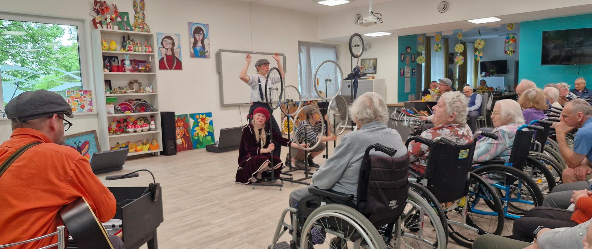 W dużej przestronnej sali trzy dziewczyny w strojach scenicznych występują przed grupą seniorów. Przed dziewczynami jest pięć kółek rowerowych na stojakach. Z przodu i z boku widać osoby, głównie starsze siedzące na fotelach lub wózkach inwalidzkich. Po lewej stronie siedzi mężczyzna w pomarańczowej koszuli i gra na gitarze. Na ścianach widać kolorowe obrazy, regał z kolorowymi ozdobami, telewizory, okna.