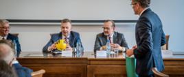 Minister Wieczorek i mężczyzna w garniturze siedzą za drewnianym stołem, minister trzyma w rękach małą żółtą piłkę.
