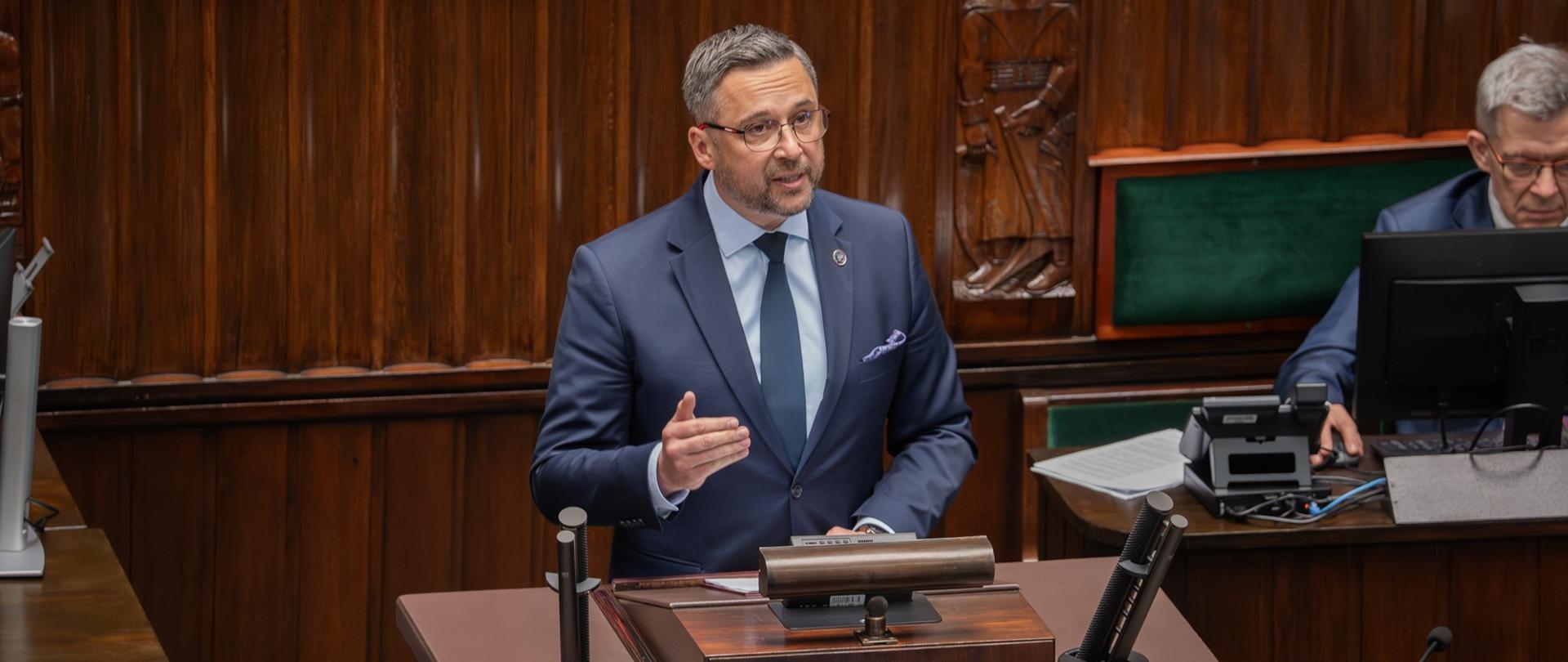 Minister Kulasek stoi za drewnianą sejmową mównicą.