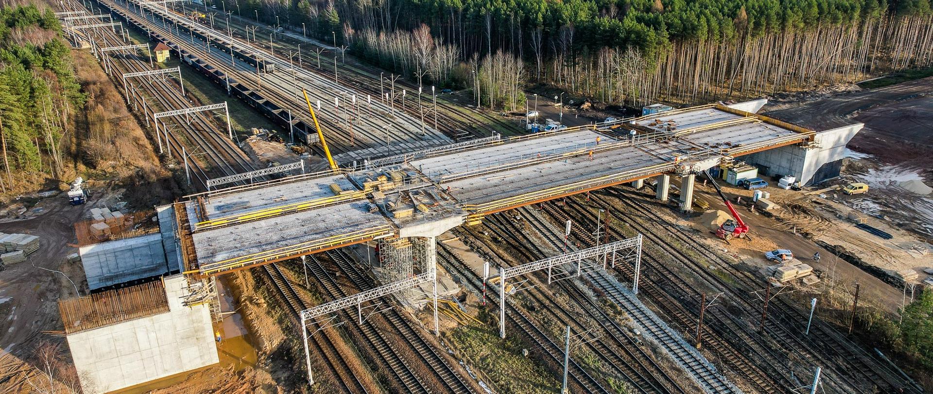 Wiadukt drogowy w budowie. Widoczne szalunki pomostu wiaduktu, oraz pracownicy i maszyny budowlane wspomagające budowę obiektu.