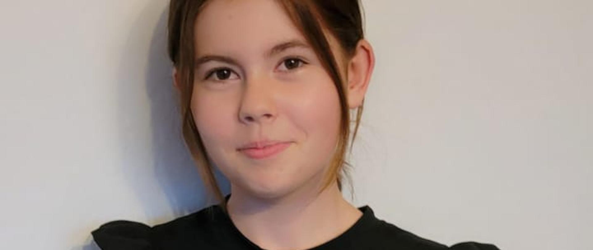 Dziewczynka ubrana na czarno stoi trzymając przed sobą w rękach dyplom.