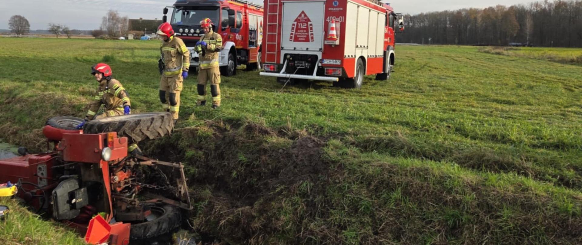 Zdjęcie przedstawia strażaków podczas ratowaniu osoby przegniecionej przez ciągnik rolniczy.
W tle samochody strażackie, pojazdy i pole.
