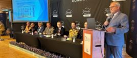 Starszy mężczyzna w szarym garniturze i okularach przemawia do mikrofonu, stojąc przy drewnianej mównicy z logo Związku Powiatów Polskich. W tle, za długim stołem prezydialnym, siedzi grupa oficjalnych gości, w tym umundurowani funkcjonariusze Państwowej Straży Pożarnej oraz cywile. Za nimi widnieje ciemna ścianka reklamowa z napisami „GÓRSKI HOTEL & SPA *** ŁÓDZKIE”.