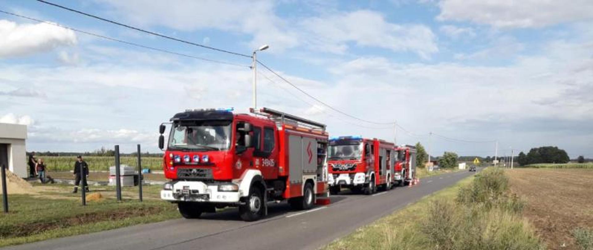 Na zdjęciu widzimy trzy wozy strażackie ustawione jeden za drugim na drodze asfaltowej. 