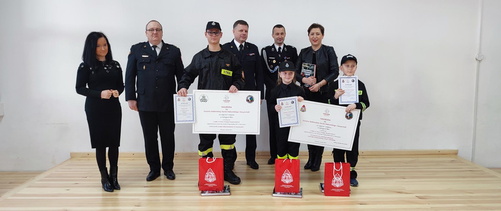 Na zdjęciu kobiety ubrane w czarne stroje, strażacy w mundurach wyjściowych koloru granatowego wraz z dziećmi ubranymi w mundury koszarowe koloru czarnego. Dzieci trzymają dyplomy i nagrody rzeczowe w torebkach koloru czerwonego. 