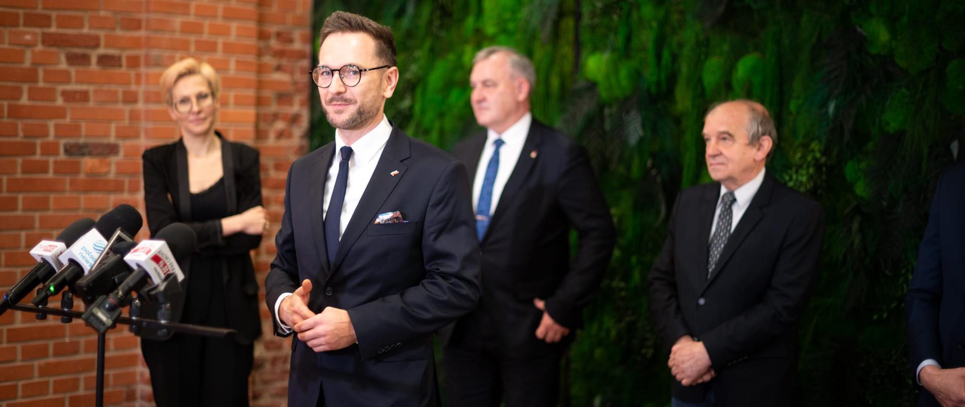 Minister Waldemar Buda przemawia na konferencji prasowej dotyczącej Technikum Automatyki i Robotyki. W tle widać dwóch mężczyzn i jedną kobieta. 