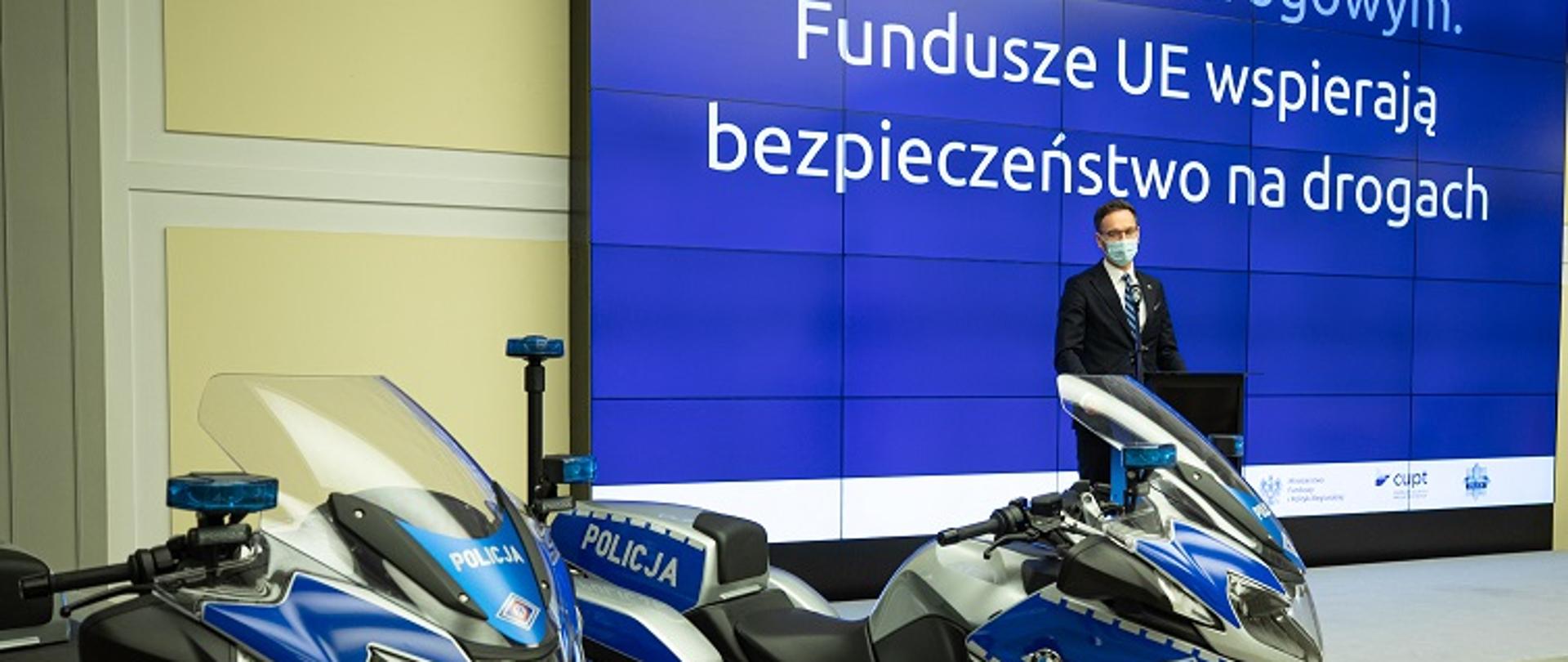 Na zdjęciu dwa motocykle oznaczone jako policyjne, za nimi stoi minister Waldemar Buda. Na ekranie napis: Fundusze UE wspierają bezpieczeństwo na drogach