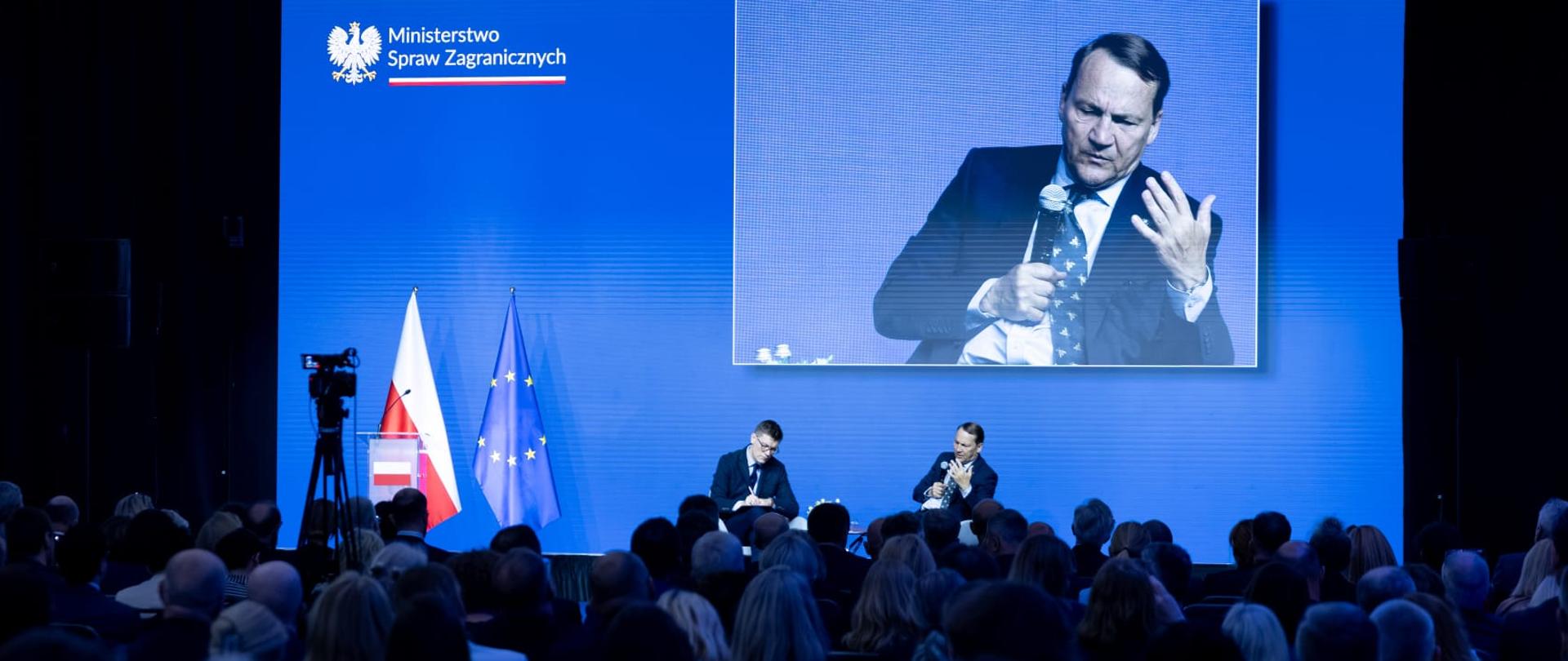 Wicepremier Sikorski na scenie bierze udział w panelu i żywo gestykuluje. Nad nim niebieskie tło z ekranem, na którym widoczny jest Wicepremier Sikorski. Przed nim widać siedzącą widownię.