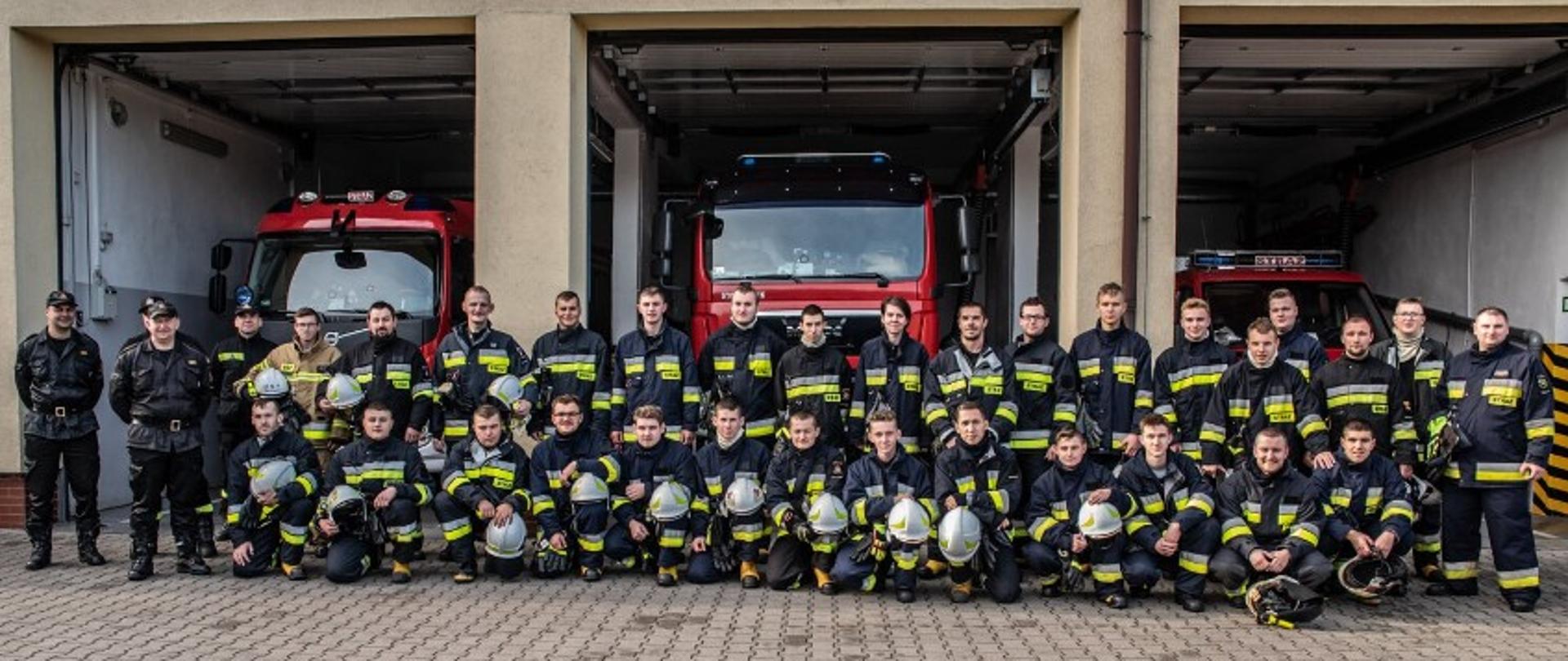 Zdjęcie grupowe druhów uczestniczących w Szkoleniu Podstawowym Strażaków Ratowników Ochotniczych Straży Pożarnych na tle samochodów pożarniczych. Strażacy w ubraniach bojowych stoją w szyku. W tle widać pojazdy pożarnicze zaparwkowane w garazach. 