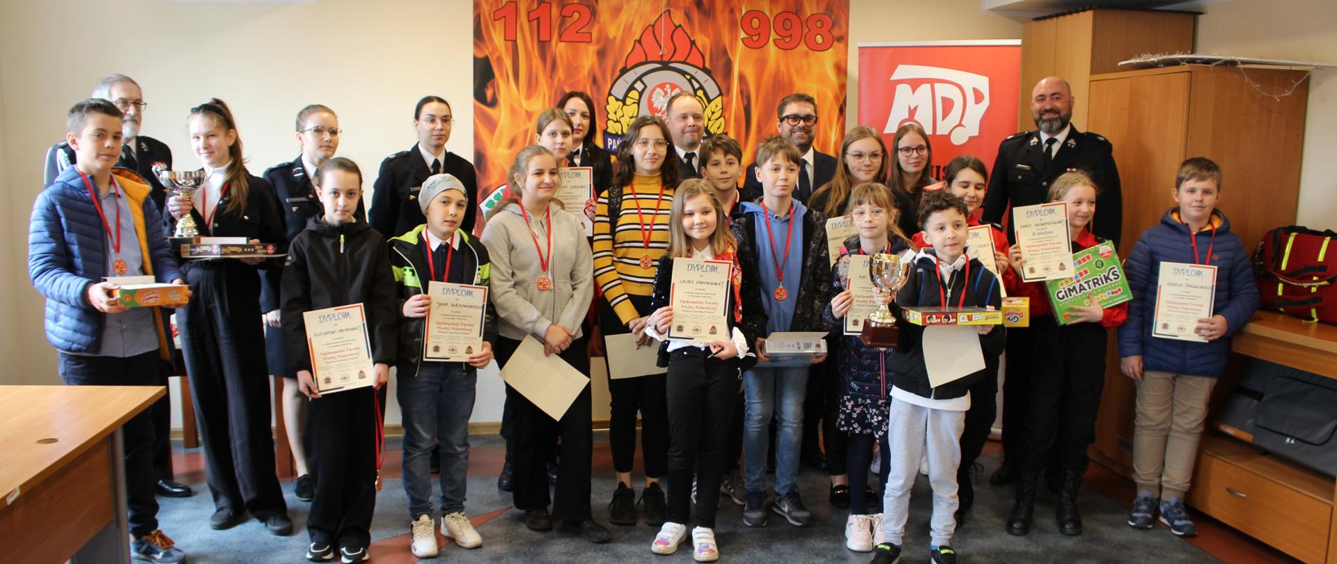 Na świetlicy stoi grupka dzieci i młodzieży z organizatorami turnieju. dzieci z dyplomami. Z tytuł na ścianie logo PSP i numery 998 i 112