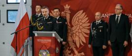 Komendant powiatowy przemawia do zaproszonych gości oraz pracowników komendy po odczytaniu aktu powołania na stanowisko jego zastępcy brygadiera Bartosza Debernego. W tle komendanci oraz zaproszeni goście.