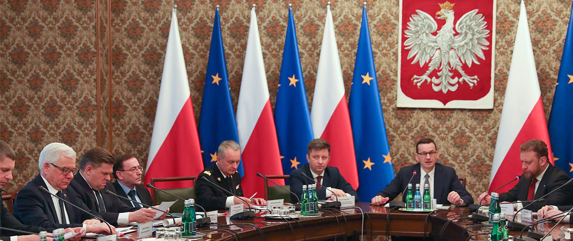 Posiedzenie Rządowego Zespołu Zarządzania Kryzysowego w KPRM . Na zdjęciu widać m.in. Mateusza Morawieckiego premiera, Łukasza Szumowskiego ministra zdrowia i Mariusza Kamińskiego ministra SWiA. Posiedzenie odbywa się w sali Kościuszkowskiej KPRM. Wszyscy siedzą przy owalnym stole.