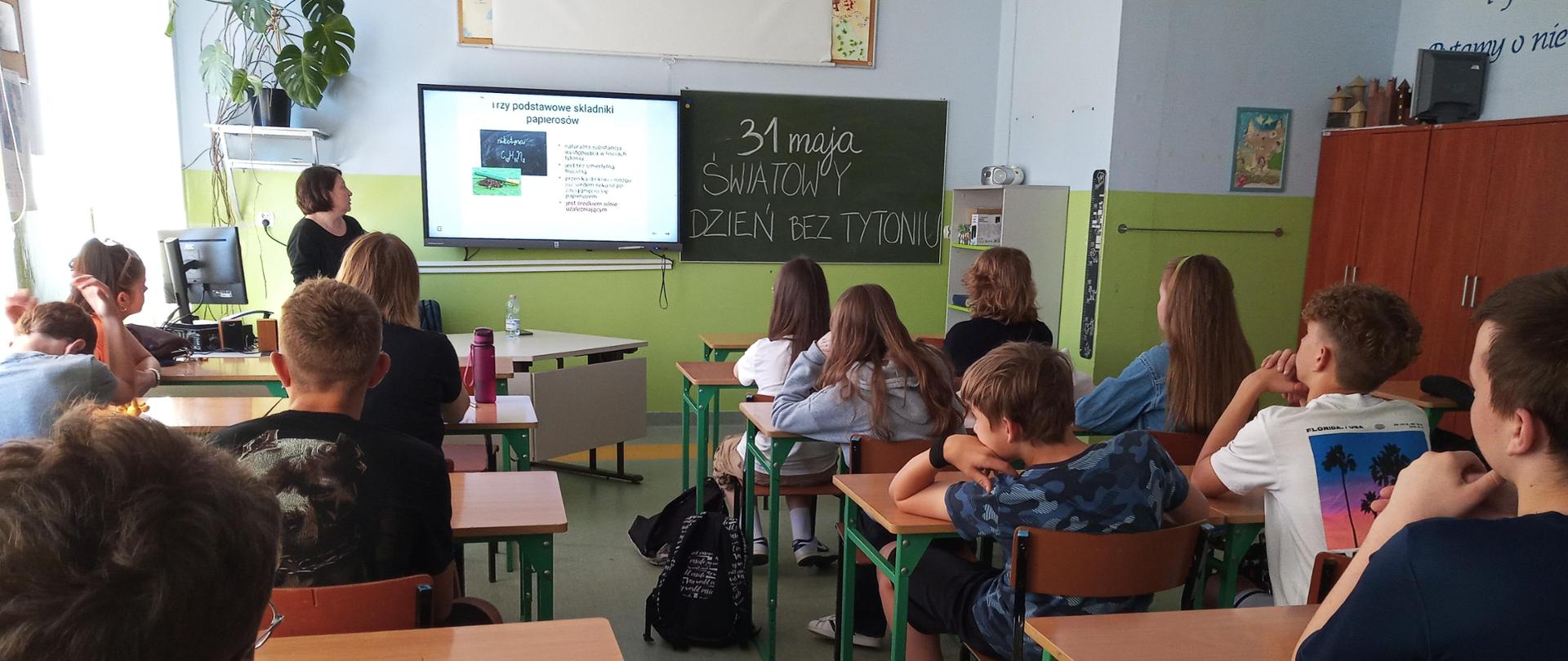 Liczna grupa uczniów (chłopców i dziewcząt) siedząca w ławkach w klasie szkolnej. Młodzież zwrócona w kierunku prelegentki oraz tablicy, na której widnieje napis „31 maja Światowy Dzień bez Tytoniu”. Obok tablicy duży ekran, na którym wyświetlony jest slajd prezentacji nt. składu dymu tytoniowego.