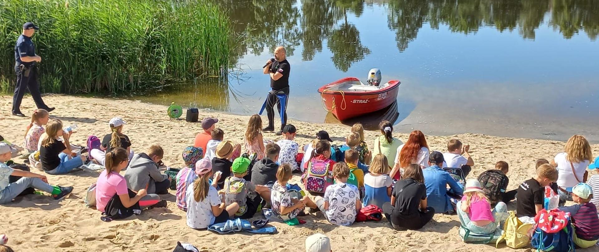 Zdjęcie przedstawia dzieci siedzące na plaży przy jeziorze i strażaka przeprowadzającego instruktaż. Obok stoi czerwona łódka
