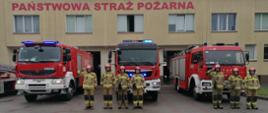 siedmiu funkcjonariuszy w mundurach na tle wozów strażackich, wozy strażackie znajdują się na tle budynku strażnicy KP PSP 