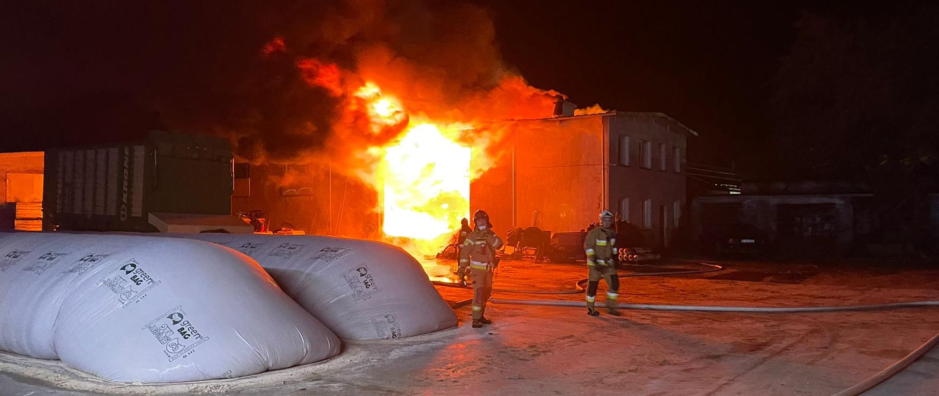 Na zdjęciu zrobionym w porze nocnej widocznych jest trzech strażaków w ubrania specjalne koloru piaskowego walczących z płomieniami, które pochłonęły budynek gospodarczy na terenie gospodarstwa rolnego. Płomienie intensywnie wychodzą z wnętrza pomieszczenia na zdjęciu widać równie przyczepę transportową, dwie pryzmy osłonięte biała folią plastikową oraz rozciągnięte na betonowym placu węże strażackie.