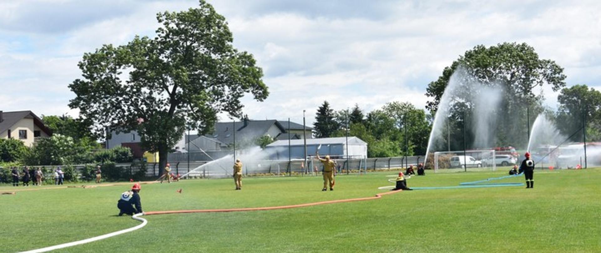 Obraz przedstawia szeroki widok na otwarte, zielone boisko sportowe, gdzie odbywają się zawody strażackie. Na pierwszym planie, na trawie z białymi liniami, widać strażaków w beżowych kombinezonach, którzy obsługują węże strażackie. Z niektórych węży tryskają potężne strumienie wody, tworząc wysokie fontanny, szczególnie po prawej stronie obrazu. W tle widoczne są budynki mieszkalne i bujne drzewa. Na horyzoncie po prawej stronie, za fontannami wody, można dostrzec fragmenty czerwonych wozów strażackich. Niebo jest częściowo zachmurzone, ale dzień wydaje się być jasny.