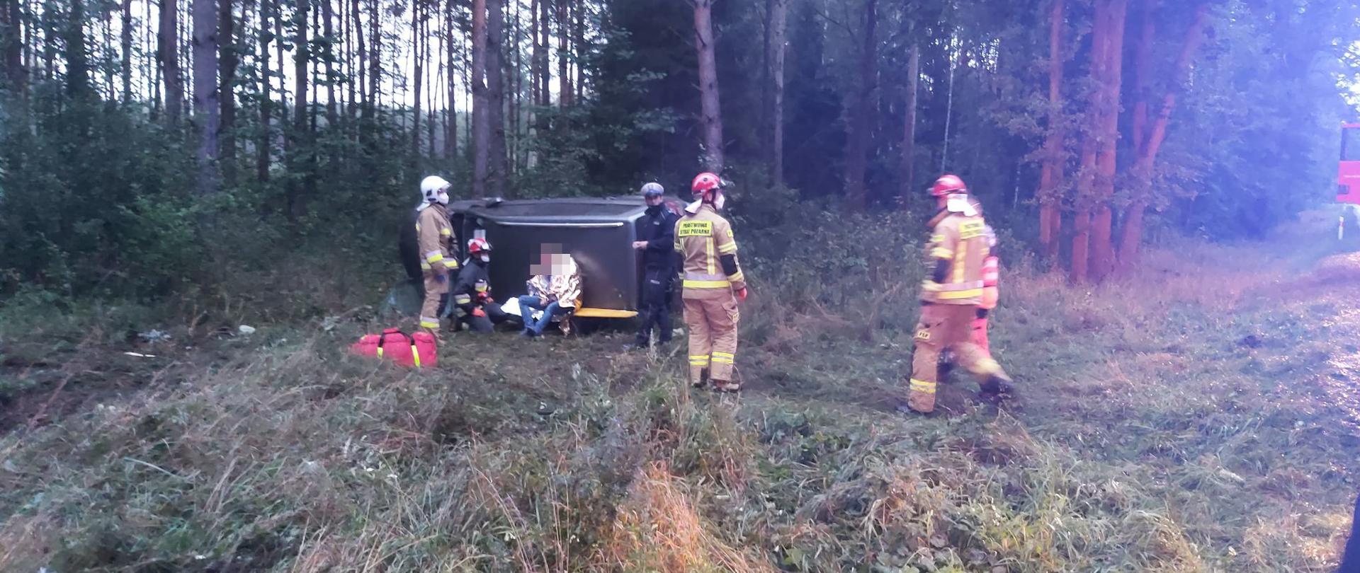 Na tle lasu na lewym boku leży samochód osobowy koloru ciemnego. O dach pojazdu oparty plecami jest uczestnik wypadku. Siedzi na desce ortopedycznej, owinięty kocem termicznym. Twarz mężczyzny zanonimizowana. Przy poszkodowanym klęczy strażak w czerwonym hełmie, obok stoją dwaj inni strażacy i policjant. Z prawej strony do poszkodowanego podchodzi kolejny strażak wraz z ratownikiem medycznym. Przed pojazdem stoi także czerwona torba ze sprzętem ratownictwa medycznego.