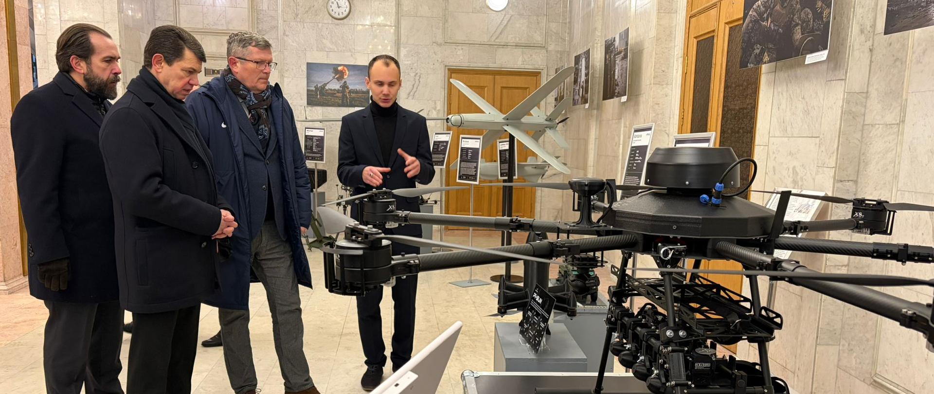 A group of men observes an unmanned aerial vehicles (drones) displayed in an exhibition hall. One presenter explains the construction elements of the device, with additional exhibits and informational panels visible in the background.