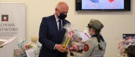 Wojewoda wręcza wygraną stojącej przed nim harcerki ubraną w mundur. Dziewczynka stoi tyłem do obiektywu. W tle stół zastawiony nagrodami oraz ekran na którym wyświetlają sie zwycięskie prace 