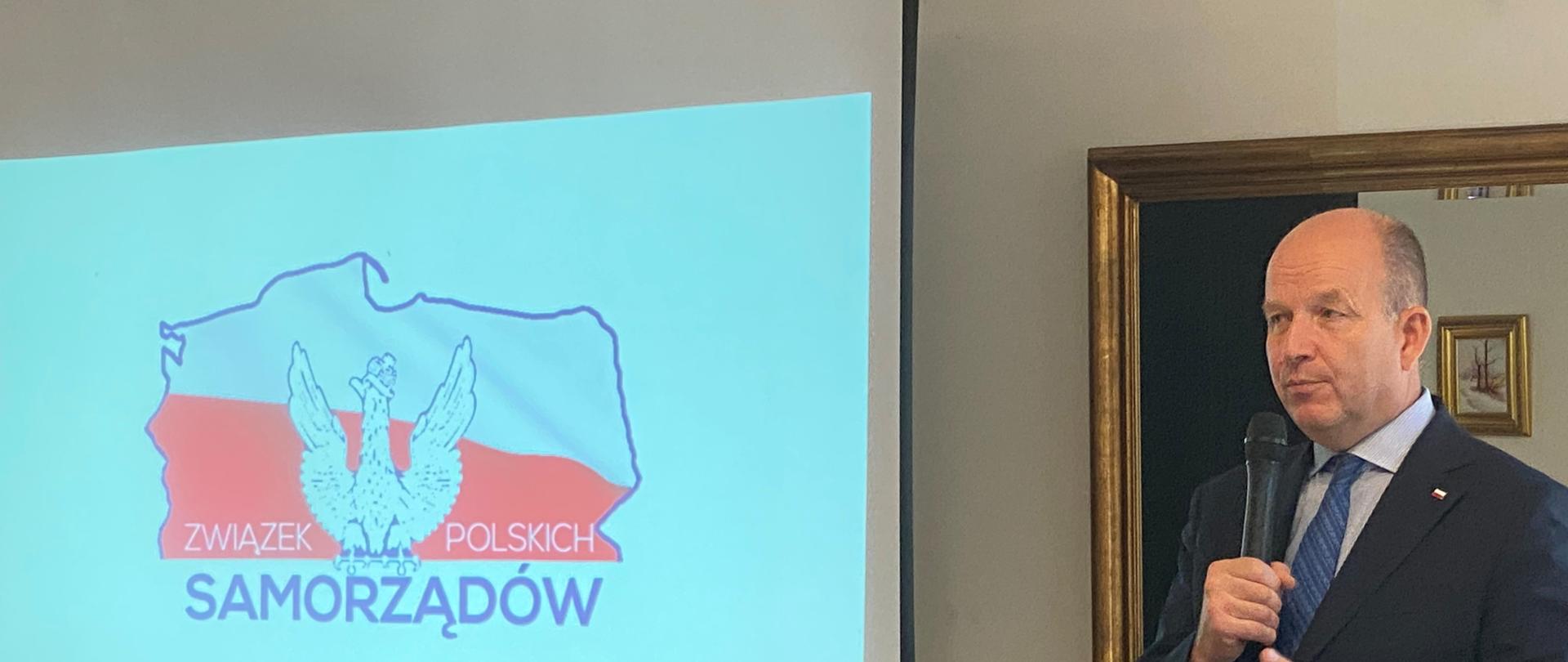 Na zdjęciu znajduje się ubrany w ciemnogranatowy garnitur, błękitną koszulę i niebieski krawat mężczyznę, który jest w trakcie wystąpienia publicznego. Mężczyzna ten trzyma w prawym ręku mikrofon. W tle, po prawej stronie mężczyzny znajduje się ekran, na którym wyświetlone jest logo Związku Polskich Samorządów. 