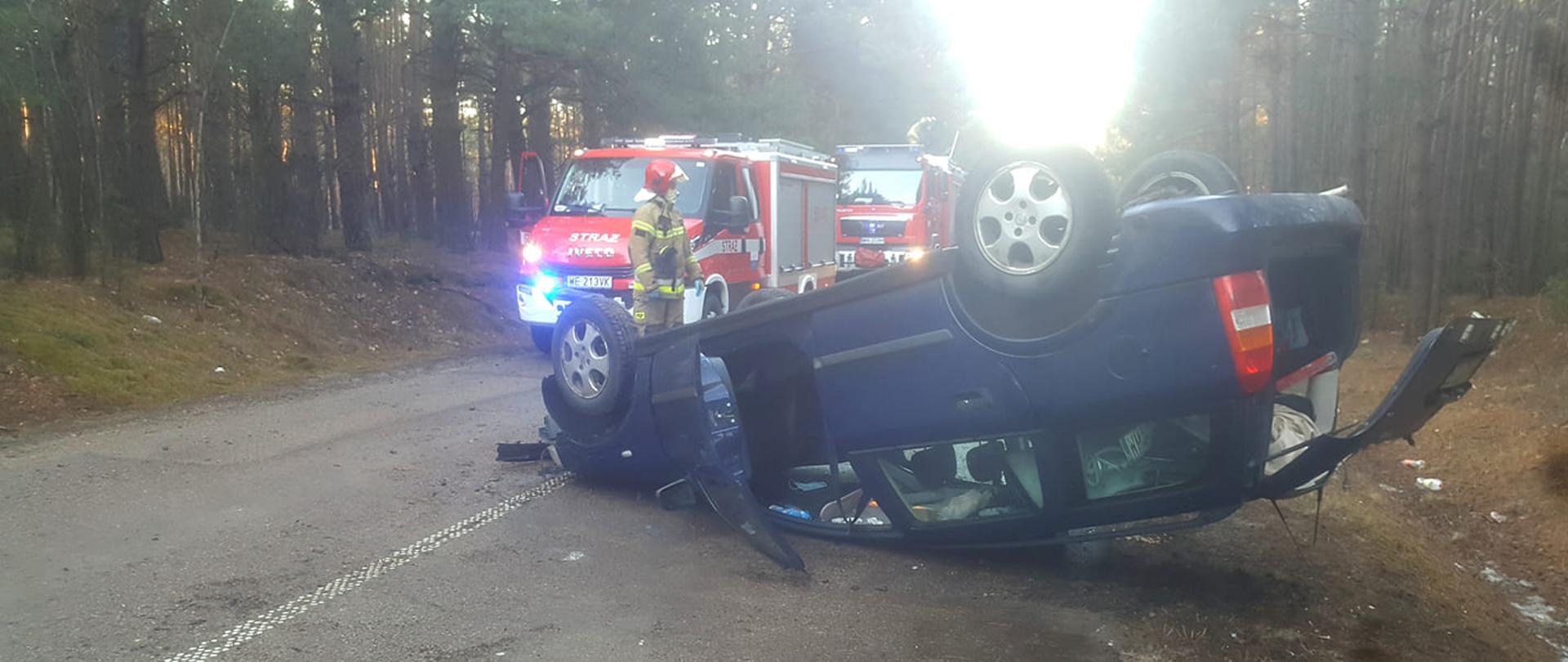 Samochód osobowy leży na dachu po wypadku na jednym pasie drogi. W tle samochody strażackie.