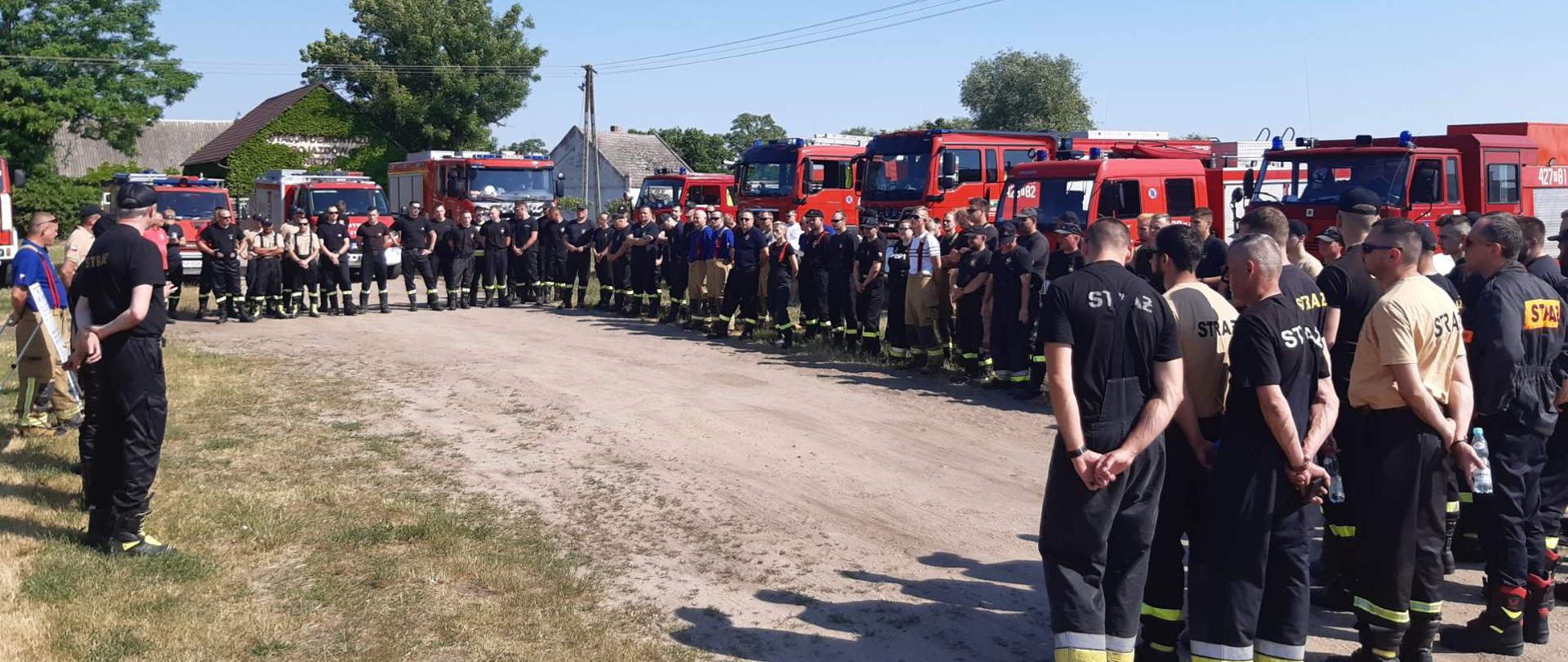 Zdjęcie grupowe uczestników ćwiczeń Integracja 2021 - odprawa przed ćwiczeniami. Grupa strażaków w ciemnych ubraniach, w tle czerwone samochody pożarnicze.